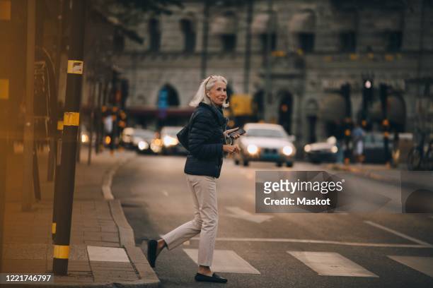 senior woman looking away while crossing road in city - variable schärfentiefe stadt stock-fotos und bilder