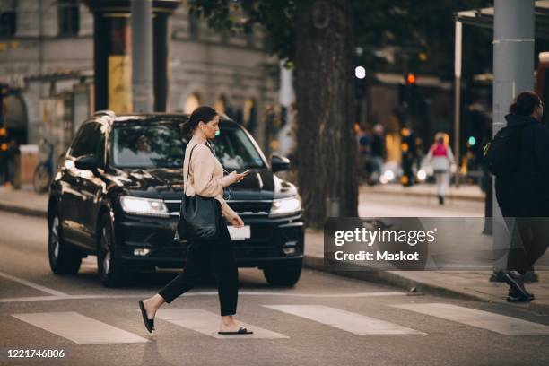 young woman using phone while walking on zebra crossing in city - oversteken stockfoto's en -beelden