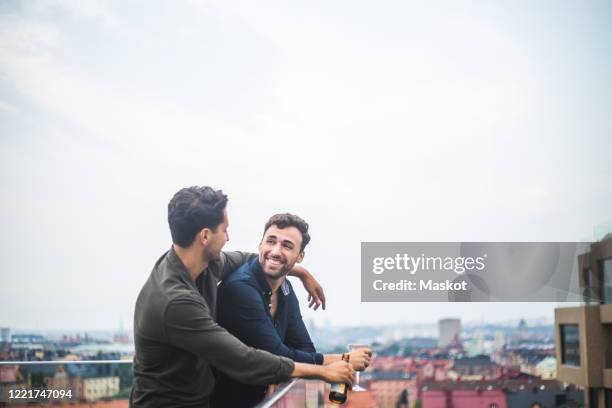 happy man looking at male friend with drink while leaning over glass railing on rooftop against sky - railings stock pictures, royalty-free photos & images