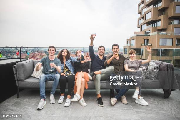 portrait of smiling male and female friends with drinks sitting on sofa at building terrace for social gathering - group on couch stock-fotos und bilder