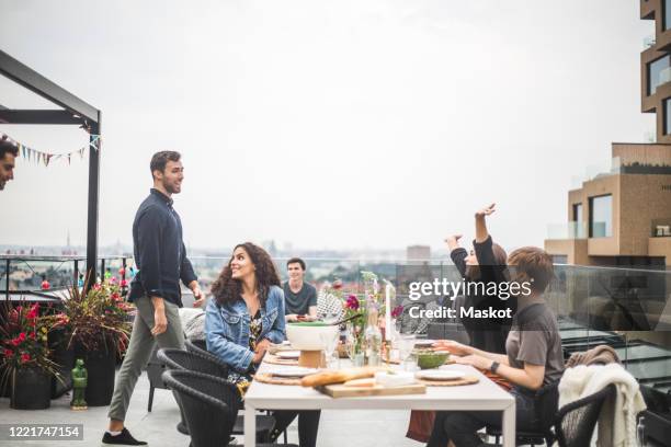 young female with arms raised greeting friends while sitting by table on terrace - rooftop party stock pictures, royalty-free photos & images