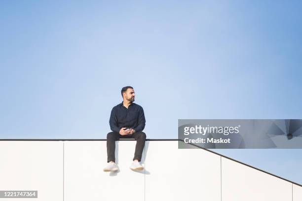 contemplating man looking away while sitting on built structure against blue sky - sitzen stock-fotos und bilder
