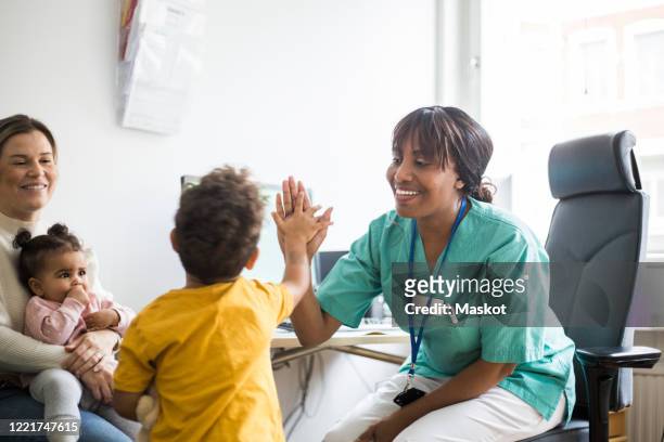 smiling pediatrician giving high-five to boy while mother looking affectionately in clinic - nurse child stockfoto's en -beelden