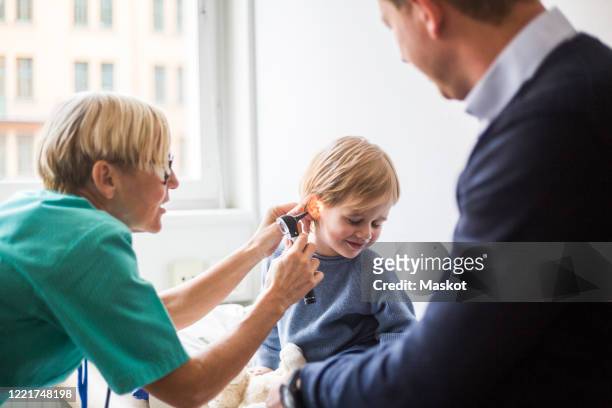 female doctor examining boy's ear with otoscope while sitting by father in hospital - otoscope bildbanksfoton och bilder