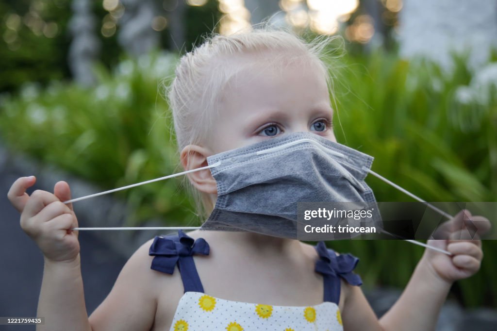 Little toddler girl trying to put medical protective mask. Candid outdoor portrait of child with medical mask. Corona virus outbreak or air pollution concept.