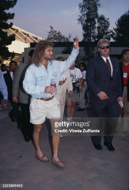 American tennis player Andre Agassi wearing a denim shirt and white shorts, holding a bottle in one hand while waving with the other, circa 1993.