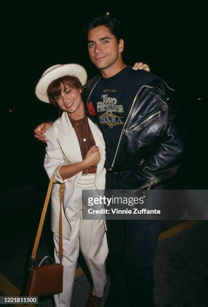 American singer and dancer Paula Abdul, wearing a white outfit with a white wide-brim hat, with American actor John Stamos, wearing a black leather...