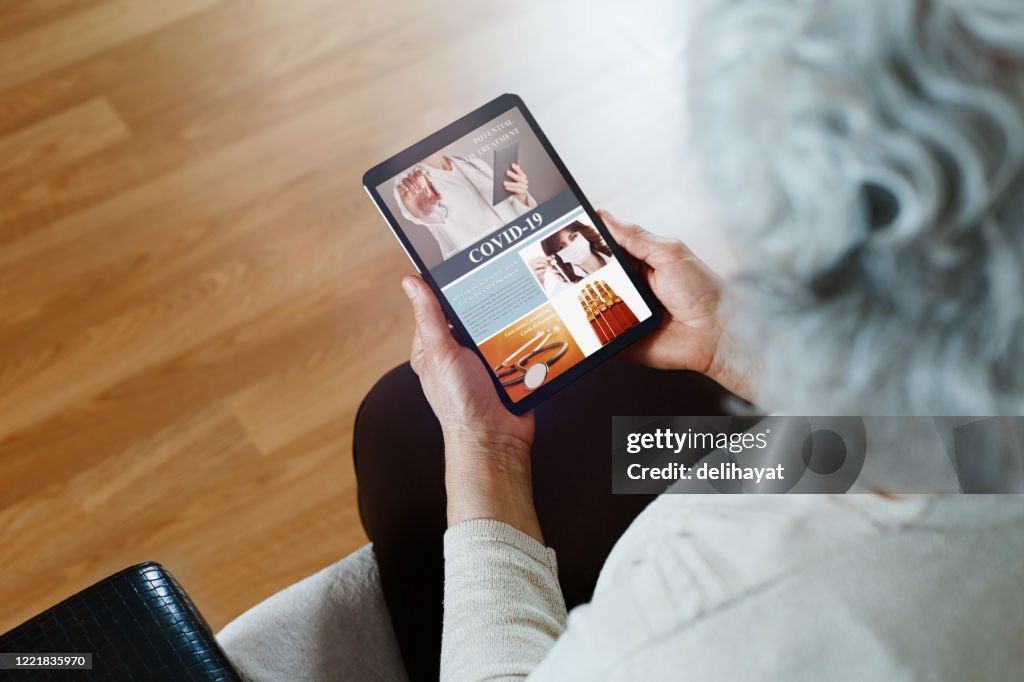 Unrecognizable elderly woman with gray hair is sitting on sofa in her living room and reading about Covid-19 new treatments and news on the digital tablet
