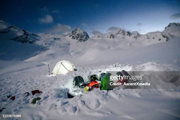 friends relax near illuminated tent in a snowy backcountry - woman snow outside night stock pictures, royalty-free photos & images