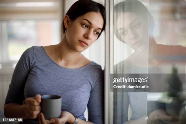 portrait of sad young woman looking through the window at home - looking through window covid stock pictures, royalty-free photos & images