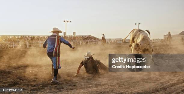 een bullrider concurreren in een bull riding event na wordt gegooid van een bull's back, terwijl de rodeo clown leidt de stier in een stadion vol mensen bij zonsondergang - bull riding stockfoto's en -beelden