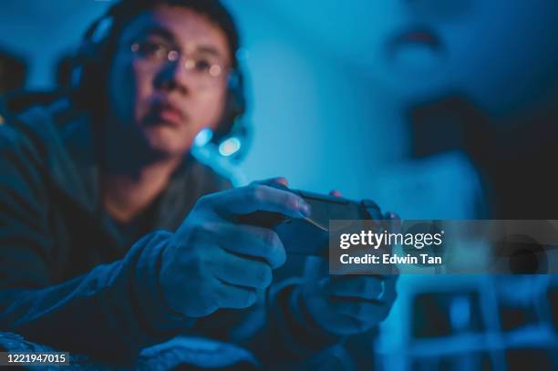 an asian chinese mid adult playing video games at home alone at night in darkness with headset and game controller sitting on sofa lying down