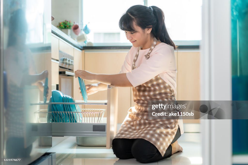 Attractive young Asian woman loading the dishwasher into cupboards at kitchen while doing cleaning at home during Staying at home using free time about their daily housekeeping routine.