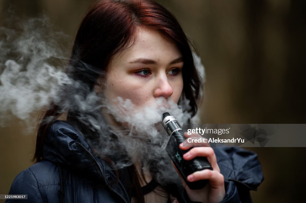 Vape teenager. Young pretty caucasian brunette girl smoking an electronic cigarette on the street in the spring. Bad habit.