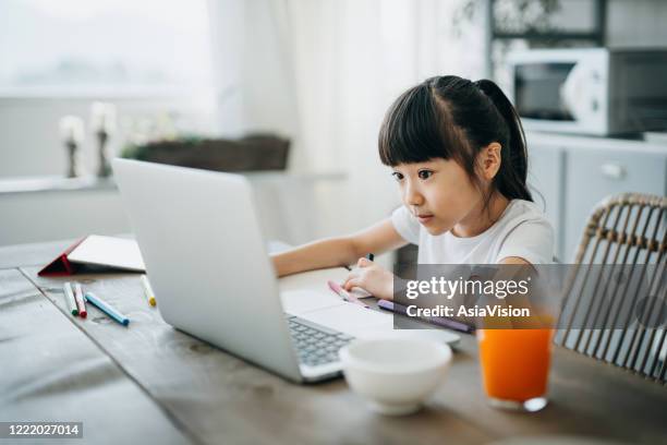 little asian girl studying from home. she is attending online school classes with laptop and digital tablet and writing notes at home - chinese tutor study stock pictures, royalty-free photos & images