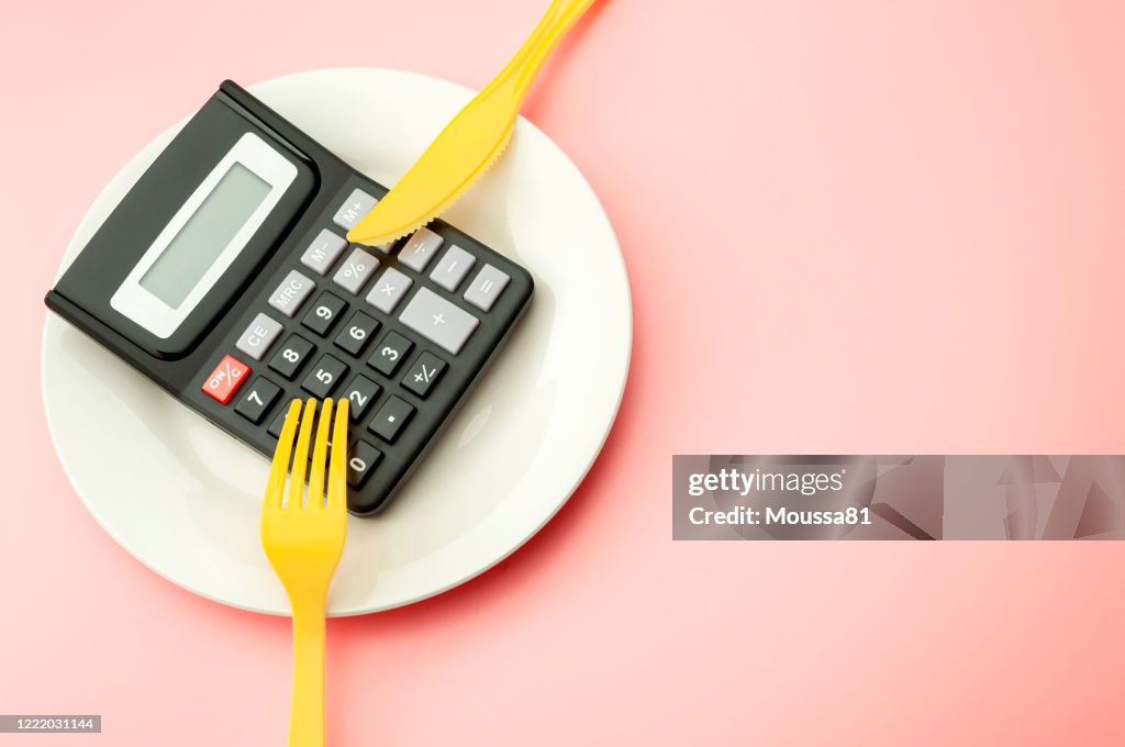 Calculate expensive food spending costs, counting calories and weight loss program concept with calculator on empty plate, yellow fork and knife isolated on pink background with copy space