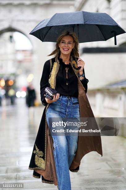 Guest wears a black top, a long coat with yellow embroidery and brown inner lining, blue jeans, during London Fashion Week Fall Winter 2020 on...