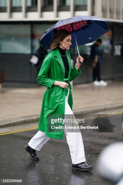 Guest wears a green shiny vinyl coat, a black turtleneck top, white pants, during London Fashion Week Fall Winter 2020 on February 16, 2020 in...