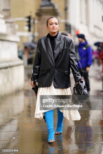 Guest wears an oversized black leather jacket, a white pleated skirt, blue long boots, a Margiela puff bag, during London Fashion Week Fall Winter...