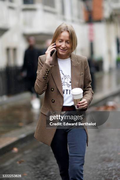 Guest wears a brown jacket, a white Tintin t-shirt, a belt, dark pants, holds a Starbucks Coffee hot drink cup, during London Fashion Week Fall...