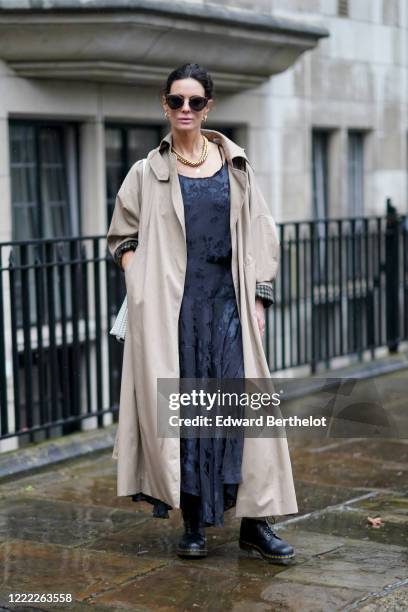 Hedvig Opshaug wears sunglasses, a golden necklace, a trench coat, a dark gray pleated dress with shiny floral print, black leather shoes, during...