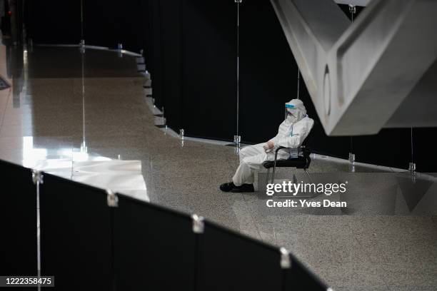 Staff worker wears a protective mask and protective suit while sitting between barriers which separate international arrivals from domestic arrivals...