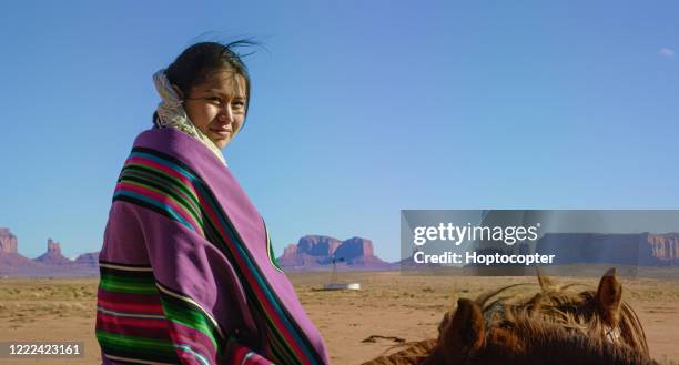 een teenaged native american girl verpakt in een traditionele navajo deken zit op haar paard en kijkt naar de camera in de monument valley desert met grote rotsformaties in de verte op een heldere, heldere dag - navajo etniciteit stockfoto's en -beelden