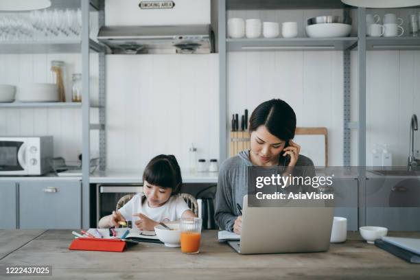 young asian mother working from home on a laptop and talking on the phone while little daughter is studying from home. she is attending online school classes with a digital tablet and doing homework at home - working mother stock pictures, royalty-free photos & images