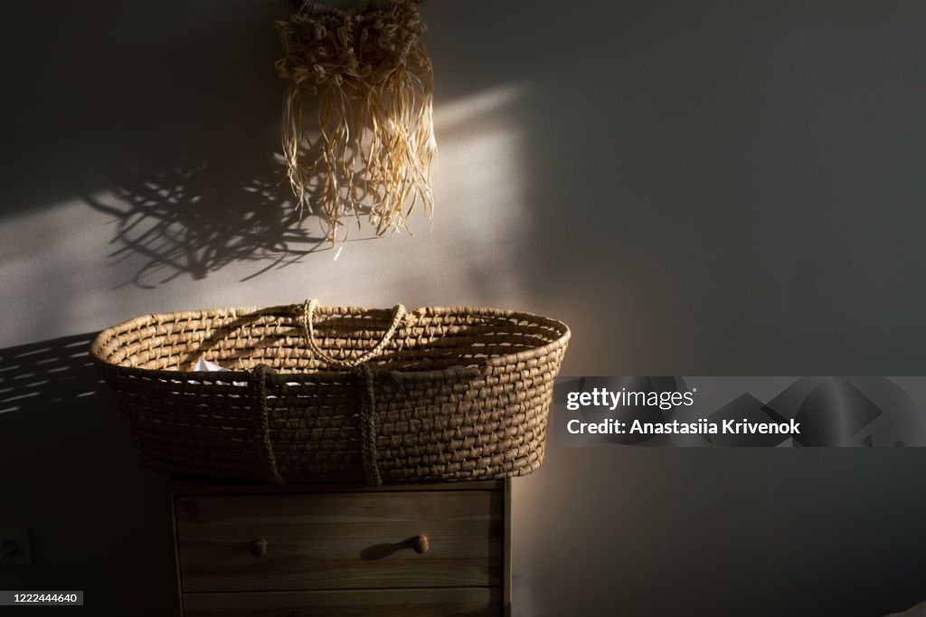 Stylish scandinavian newborn baby room with wicker bassinet on wood commode and raffia macrame panno on wall. Modern interior with beige background walls, wooden furniture and handmade macrame in sunset lights. Expecting baby concept.