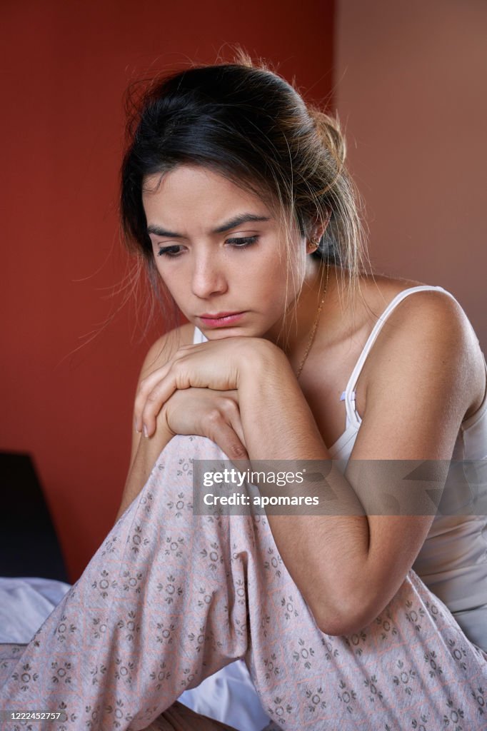 Sad and lonely Hispanic cute young woman wearing pajamas and sitting on bed