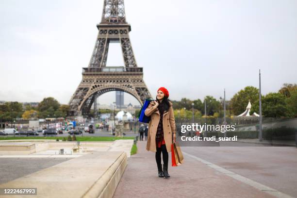 fröhliche asia frau ist zu fuß in der stadt. sie hält eine einkaufstasche in der hand und benutzt ihr smartphone. - paris millenials stock-fotos und bilder