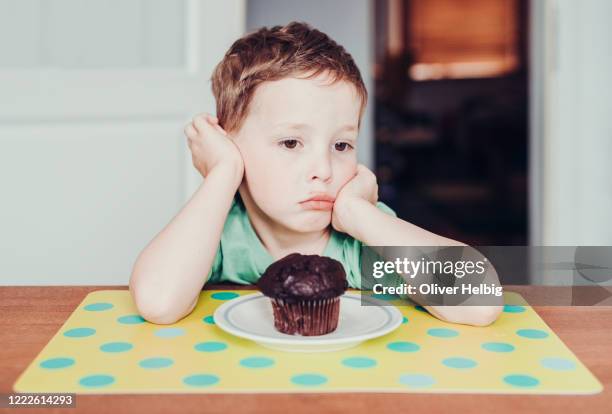 cute little boy sitting at the kitchen table. in front of him is a plate with a chocolate muffin.the little boy is making a discontented face. - unpleasant taste stock pictures, royalty-free photos & images