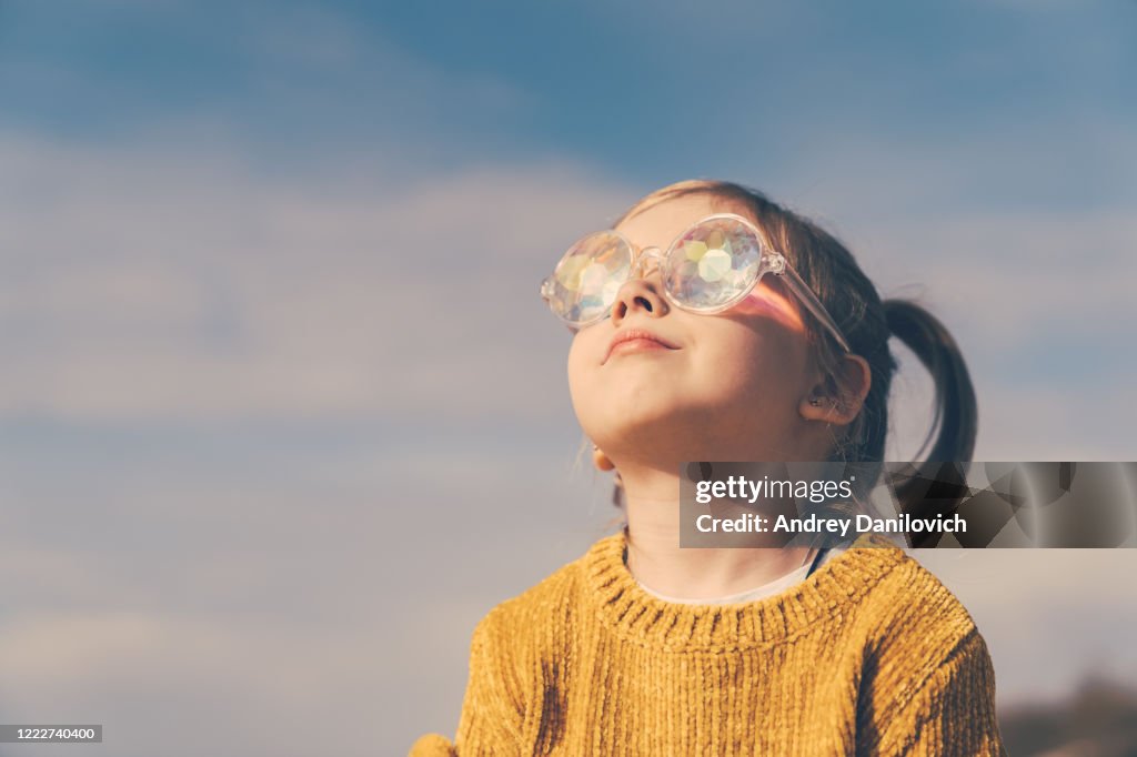 Little girl in funny kaleidoscope glasses is looking to the sky and smiling.