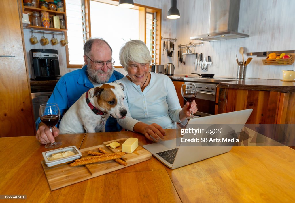 COVID-19 Stay connected. Happy senior couple with pet dog and wine video calling friends on laptop or online chatting with family celebrating easing of coronavirus restrictions lockdown. Hope concept.