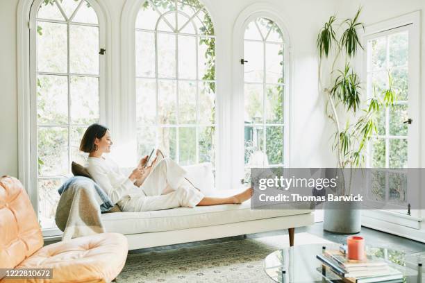 woman sitting on couch in living room reading on digital tablet - 心地よい ストックフォトと画像