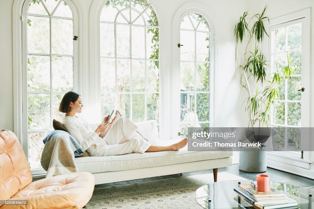 Woman sitting on couch in living room reading on digital tablet