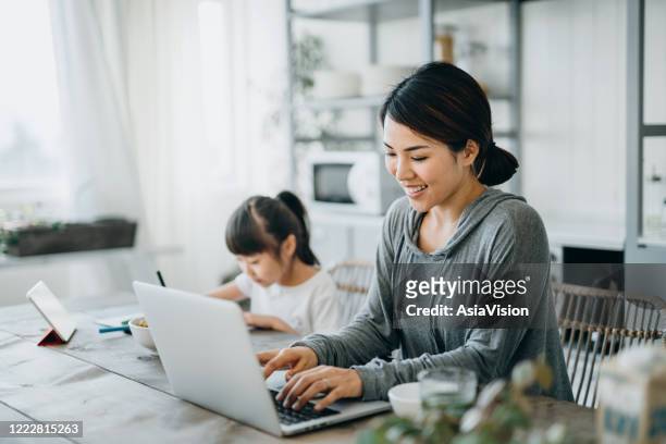 young asian mother working from home on a laptop while little daughter is studying from home. she is attending online school classes with a digital tablet and doing homework at home - working from home with kids stock pictures, royalty-free photos & images