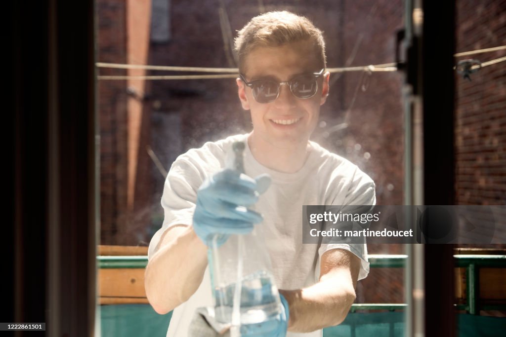 Millenial man cleaning window on city balcony in spring.