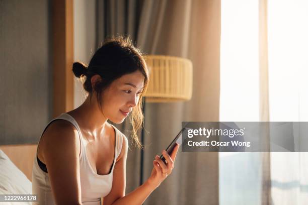 young woman using smartphone while waking up in bed - asian woman smiling sunrise stock pictures, royalty-free photos & images