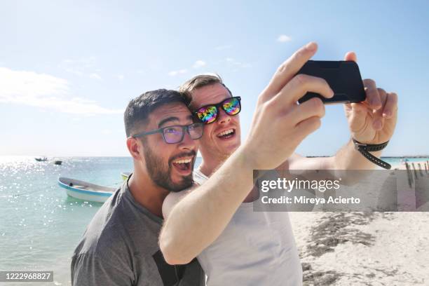 a mixed race, male couple make playful expressions for a selfie on a tropical beach during vacation. - grey boot stock-fotos und bilder