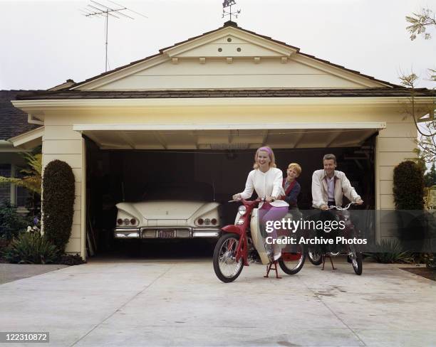 Family sitting on Honda 50 mopeds in front of garage, smiling