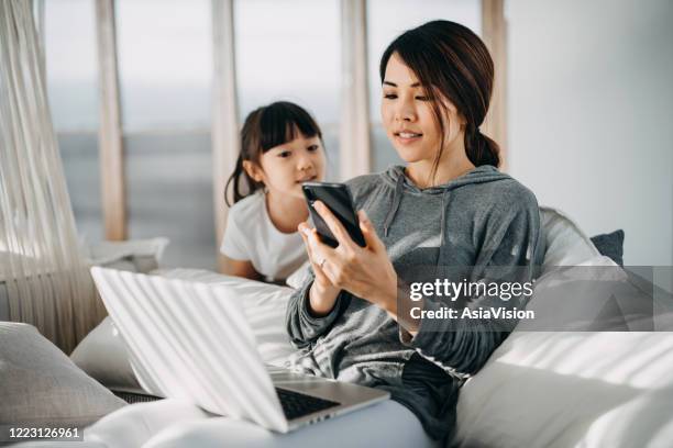 busy young asian mother sitting in bed using smartphone and working on laptop on her lap while little daughter is distracting her and seeking for attention - multitasking parent stock pictures, royalty-free photos & images