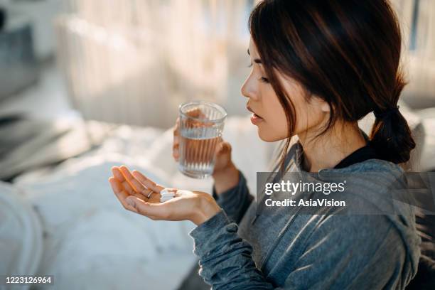 junge asiatische frau sitzt auf dem bett und fühlt sich krank, nehmen medikamente in der hand mit einem glas wasser - antibiotics stock-fotos und bilder