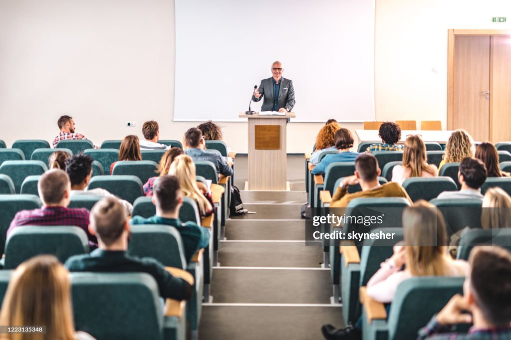 Happy mature professor giving a lecture in front of projection screen at lecture hall.