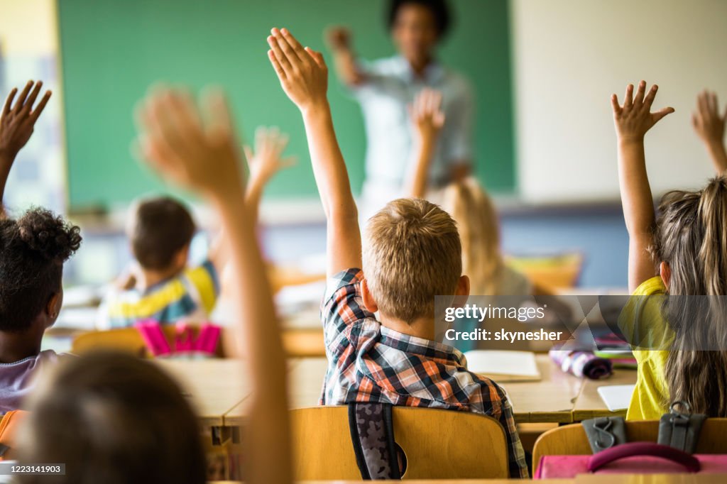Vue arrière des élèves du primaire levant la main sur une classe.