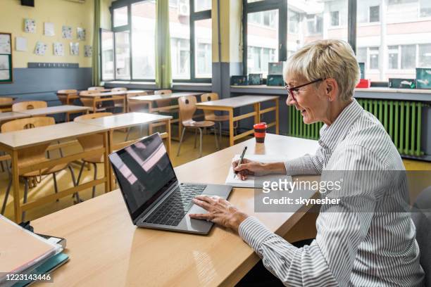 smiling senior teacher taking notes while using laptop in the classroom. - teacher stock pictures, royalty-free photos & images