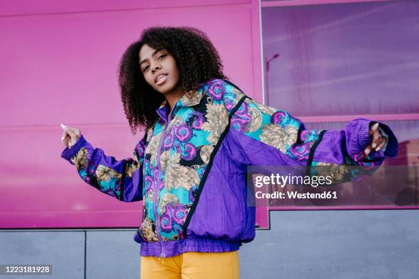 young woman with urban look dancing in front of pink glass wall - multi colored jacket stockfoto's en -beelden