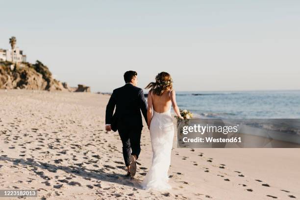rear view of happy bridal couple running at the beach - matrimoni foto e immagini stock