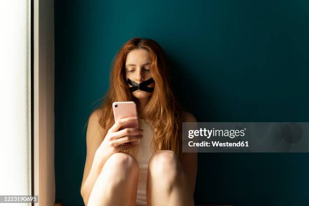 young woman with taped mouth using cell phone - censorship stockfoto's en -beelden