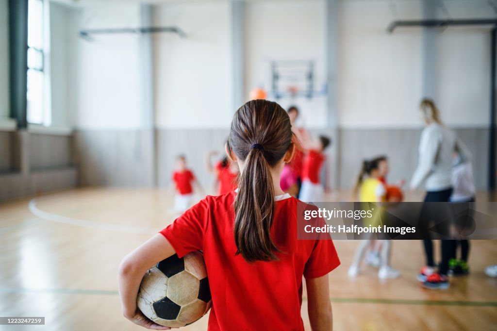 Rear view of small girl standing with ball indoors in gym class, physical education concept.